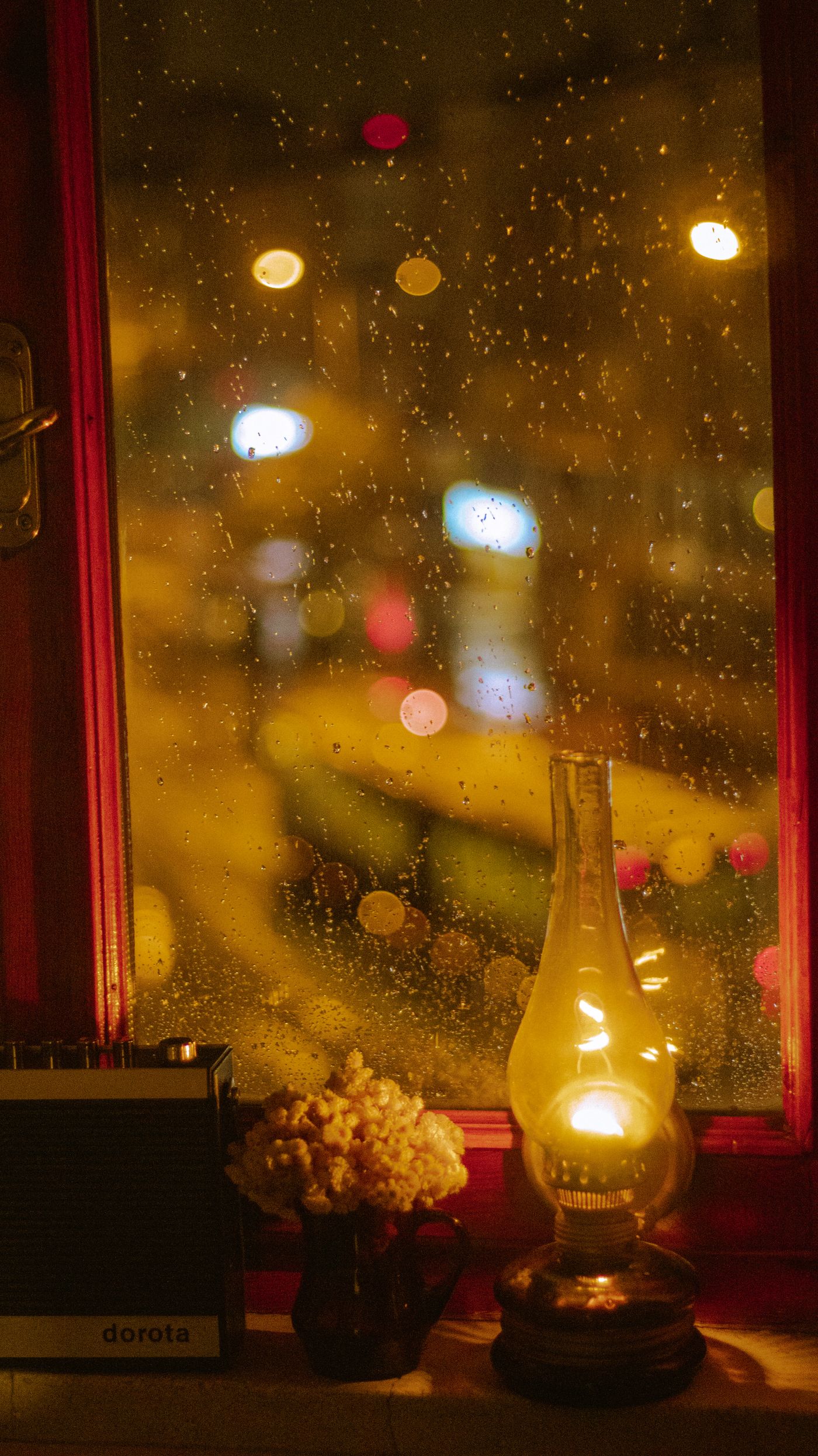 A lamp on a windowsill against rain-blurred city lights at night
